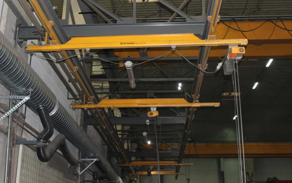 BM Makina BKB ve ALUBKB Hafif Vinç Sistemleri ile donatılmış atölye içi askılı köprü vinç hattı, tavana monte raylar üzerinde çalışan sarı gezer kirişler ve zincirli elektrikli vinçler ile üretim alanında yük taşıma çözümlerini göstermektedir.