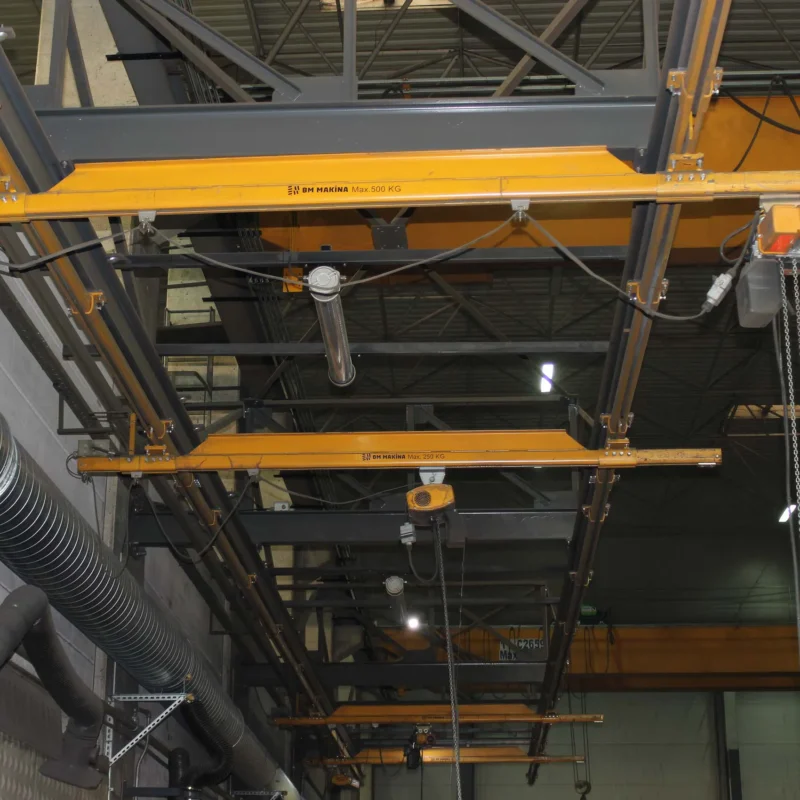BM Makina BKB ve ALUBKB Hafif Vinç Sistemleri ile donatılmış atölye içi askılı köprü vinç hattı, tavana monte raylar üzerinde çalışan sarı gezer kirişler ve zincirli elektrikli vinçler ile üretim alanında yük taşıma çözümlerini göstermektedir.