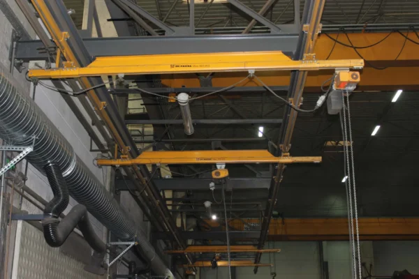BM Makina BKB ve ALUBKB Hafif Vinç Sistemleri ile donatılmış atölye içi askılı köprü vinç hattı, tavana monte raylar üzerinde çalışan sarı gezer kirişler ve zincirli elektrikli vinçler ile üretim alanında yük taşıma çözümlerini göstermektedir.